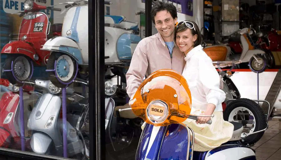 a couple standing with freshly purchased blue scooter outside of a dealership